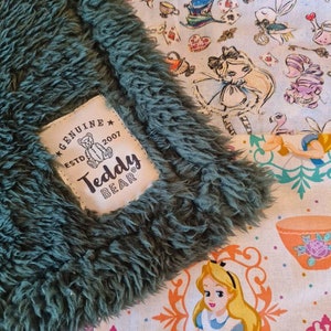 May include: A green fluffy blanket with a white label that says "Genuine Teddy Bear Est. 2007". The blanket is folded over a multi-coloured fabric with an Alice in Wonderland theme.