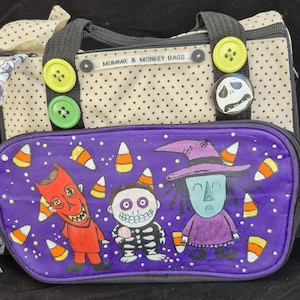 Peut inclure: Un sac fait à la main avec un panneau avant violet représentant des personnages de dessins animés et des bonbons. Le sac a un dessus crème à pois avec des poignées noires et des boutons décoratifs. Le texte "MUMMIE & MONKEY BAGS" est visible.