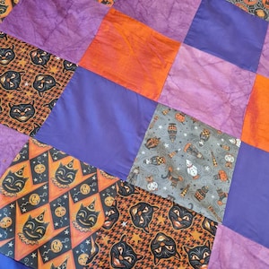 May include: A patchwork quilt with a Halloween theme. The quilt is made of various fabrics with different patterns, including black cats, pumpkins, and stars. The quilt is in shades of purple, orange, and gray.