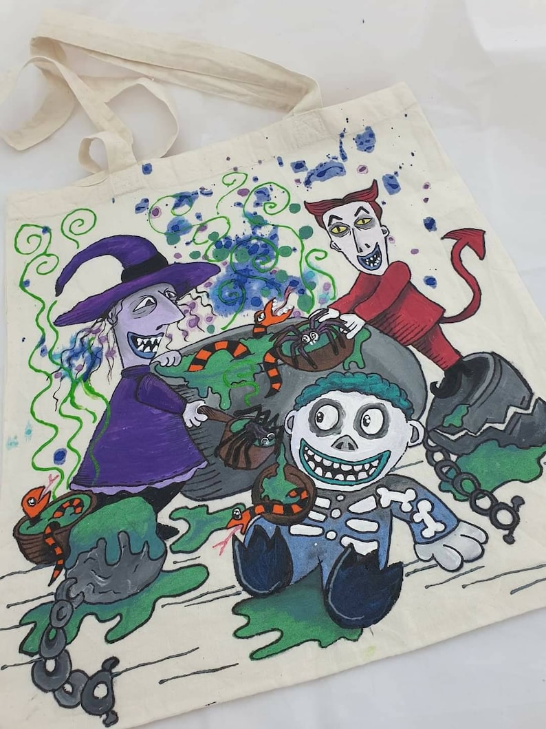 May include: A white canvas tote bag with a hand-painted design featuring characters from the Nightmare Before Christmas. The characters include Sally, Oogie Boogie, and Jack Skellington. The design is colorful and includes green, purple, red, and black.