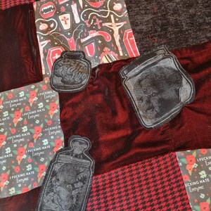 May include: Three black fabric patches featuring a design of a jar containing a human brain. The patches are sewn onto a red velvet fabric background. The background fabric has a black and red checkered pattern.