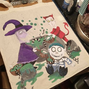 May include: A white canvas tote bag with a hand-painted design featuring characters from the Nightmare Before Christmas. The characters include Sally, Oogie Boogie, and Jack Skellington. The design is in shades of green, purple, red, and black.