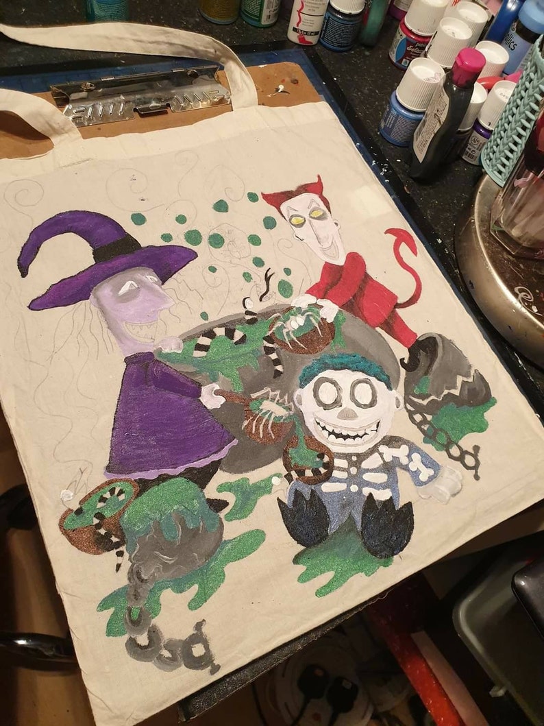May include: A white canvas tote bag with a hand-painted design featuring characters from the Nightmare Before Christmas. The characters include Sally, Oogie Boogie, and Jack Skellington. The design is in shades of green, purple, red, and black.