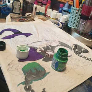 May include: A white canvas tote bag with a black and white line drawing of three witches. The bag is being painted with green, purple, and black paint. The paint is in small bottles with white lids.