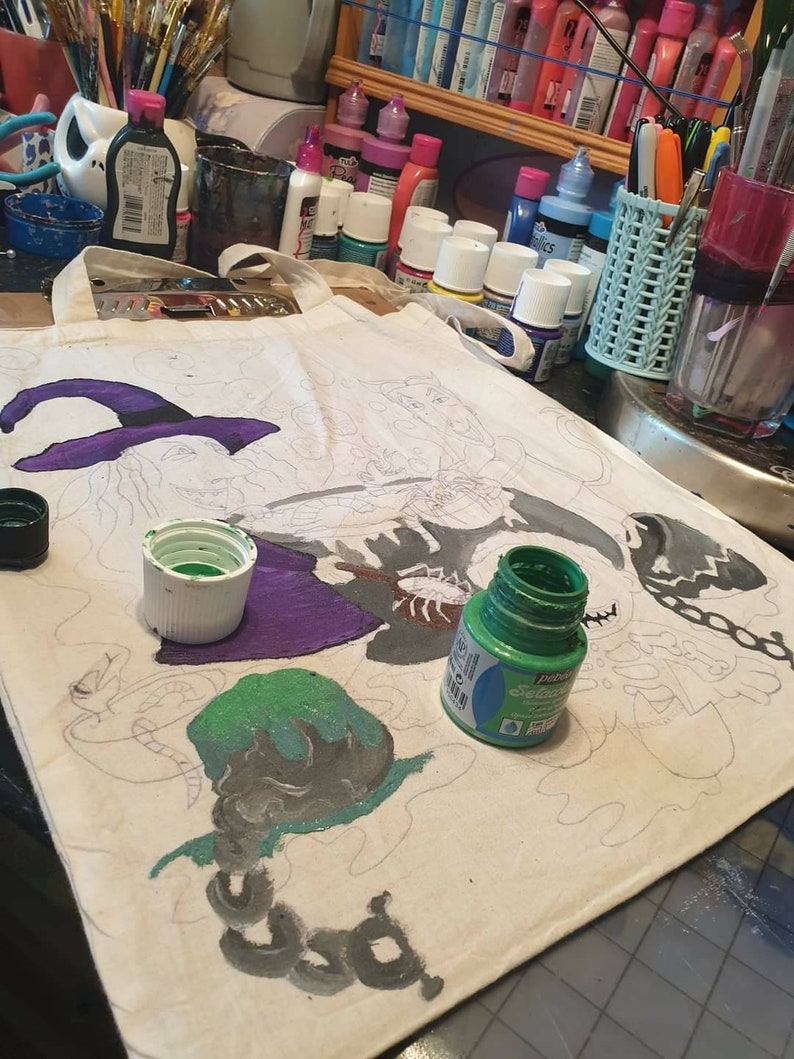 May include: A white canvas tote bag with a black and white line drawing of three witches. The bag is being painted with green, purple, and black paint. The paint is in small bottles with white lids.
