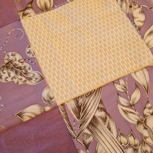 May include: A patchwork quilt with four squares. The top left square is a purple fabric with a brown floral pattern. The top right square is a yellow fabric with a woven texture. The bottom left square is a solid purple fabric. The bottom right square is a purple fabric with a brown floral pattern featuring a bird.