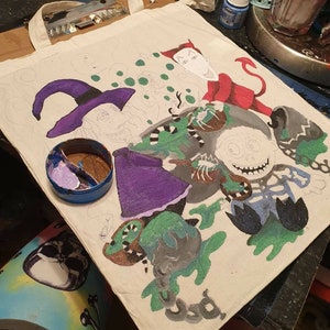 May include: A white canvas tote bag with a hand-painted design featuring a witch, a skeleton, and a devil. The design is in shades of green, purple, black, and red.