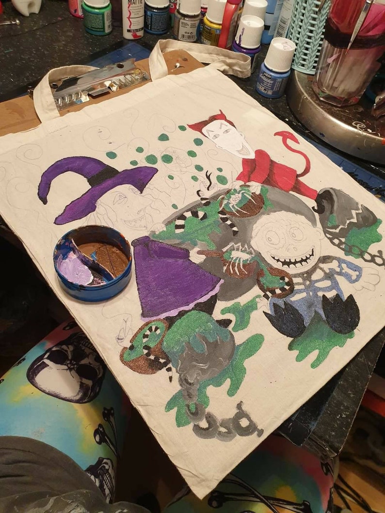 May include: A white canvas tote bag with a hand-painted design featuring a witch, a skeleton, and a devil. The design is in shades of green, purple, black, and red.