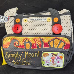 Peut inclure: Un sac fourre-tout à pois noirs et blancs avec une fermeture éclair noire et un motif noir et jaune sur le devant. Le motif représente une silhouette noire d'une maison avec un toit jaune et les mots "Simply Meant to Be". Il y a aussi plusieurs petits autocollants colorés sur le sac.