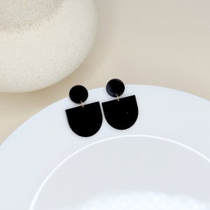 May include: A pair of black geometric earrings with a half-circle shape and a small round accent. The earrings are on a white surface.