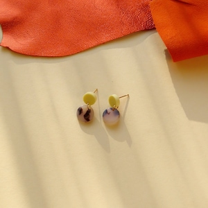 Può includere: Un paio di orecchini con un design circolare giallo e bianco. Gli orecchini hanno un piccolo cerchio giallo sopra un cerchio bianco più grande con venature nere.