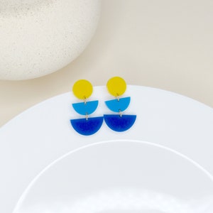 Könnte beinhalten: Ein Paar geometrische Ohrringe in Gelb, Blau und Dunkelblau. Die Ohrringe sind aus Acryl und haben eine Halbkreisform.