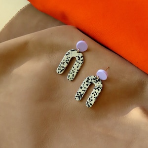 Può includere: Un paio di orecchini pendenti di colore bianco e lilla chiaro. Gli orecchini hanno la forma di un arco e un motivo maculato bianco e nero.