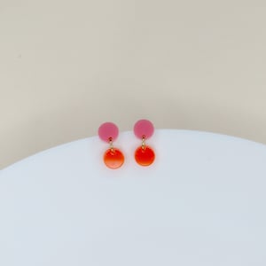 Könnte beinhalten: Ein Paar Ohrringe in Pink und Orange. Die Ohrringe bestehen aus zwei Kreisen, einem rosa und einem orangefarbenen, die durch einen goldenen Draht verbunden sind.