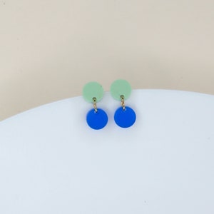 Puede incluir: Un par de pendientes con círculos de dos tonos. El círculo superior es verde claro y el círculo inferior es azul.
