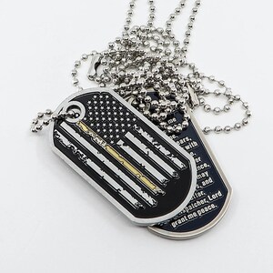 May include: Two silver dog tags on a silver chain. One tag features a black and white American flag with a thin gold line, the other tag has a black background with white text that reads "me ne ears. with e. er nce. may sand ter spatcher, Lord grant me peace."