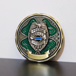 May include: A round, gold-toned police officer challenge coin with a green shamrock design. The coin features a police badge with the words "POLICE OFFICER" and "SERVE & PROTECT". A blue line is visible on the badge.