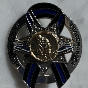 May include: A silver and black pin with a blue ribbon and a gold star in the center. The pin says "Blessed are the Peacemakers" around the outside.