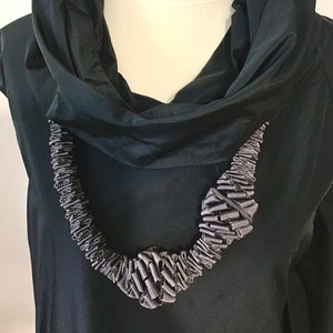 May include: A black cowl neck top with a black and white ribbon necklace. The necklace is made of multiple loops of ribbon that are twisted and knotted together.