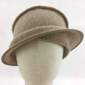 May include: A taupe-colored cloche hat with a rounded crown and a slightly downward-sloping brim. The hat has a textured, felt-like appearance and a thin, darker band around the brim's edge. The hat is displayed on a mannequin head.