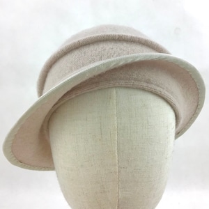 May include: A light beige cloche hat with a wide brim and a structured crown. The hat has a subtle texture and a contrasting trim around the brim. The hat is displayed on a mannequin head.