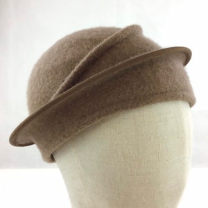 May include: A taupe-colored cloche hat with a curved brim and a decorative band. The hat is made of a soft, textured material and is displayed on a mannequin head. The hat's design is classic and elegant, suitable for various occasions.