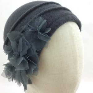 May include: A dark gray cloche hat with a decorative flower made of layered fabric and netting. The hat has a ribbed design across the top and is displayed on a mannequin head. The flower is a darker shade of gray than the hat.