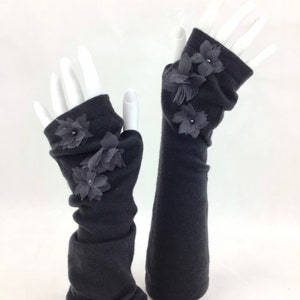 May include: A pair of dark gray fingerless gloves with decorative gray fabric flowers. The gloves are displayed on white mannequin hands against a white background. The gloves are made of a soft, knitted material.