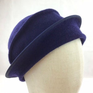 May include: A deep indigo blue cloche hat with a dark blue ribbon trim. The hat has a rounded crown and a slightly upturned brim. The hat is displayed on a mannequin head against a white background.