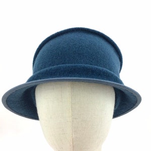 May include: A navy blue cloche hat with a rounded crown and a small brim. The hat is made of a soft, felt-like material and features a darker blue trim around the brim. The hat is displayed on a mannequin head against a white background.