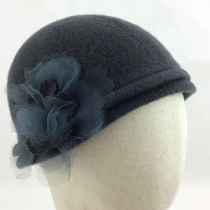 May include: A dark gray cloche hat with a decorative blue-gray flower and netting. The hat has a rounded crown and a small brim. The flower is made of layered fabric and netting, adding a touch of elegance to the design.