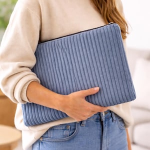 Könnte beinhalten: Eine hellblaue Cord-Clutch mit schwarzem Reißverschluss. Die rechteckige Tasche hat vertikale Rippen und wird von einer Person gehalten, die einen cremefarbenen Pullover und eine blaue Jeans trägt.