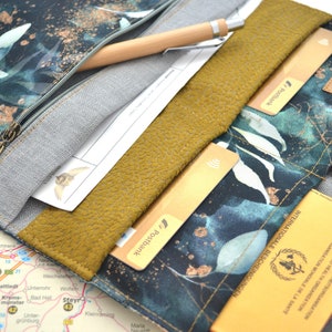 Könnte beinhalten: Eine dunkelblaue Reisegeldbörse mit einem goldenen und weißen Blumenmuster. Die Geldbörse hat mehrere Fächer für Karten, Bargeld und Dokumente. Sie hat auch ein Reißverschlussfach für einen Stift. Die Geldbörse ist geöffnet und zeigt einen Reisepass, Kreditkarten und andere Reisedokumente.