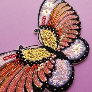 May include: A handcrafted butterfly design, intricately beaded with a black outline. The wings display a vibrant array of pink, orange, gold, and white beads and sequins. The butterfly is set against a soft purple backdrop, highlighting the detail.