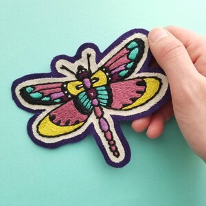 May include: Embroidered dragonfly patch with a purple border. The dragonfly features wings in shades of pink, yellow, teal, and black. The patch is held against a light blue background.