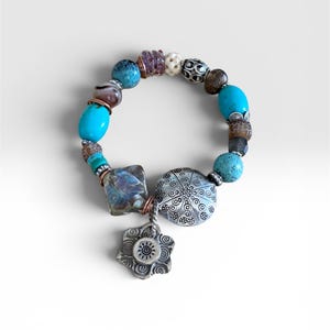 Op de afbeelding: Een kralenarmband met diverse turquoise, bruine en paarse kralen afgewisseld met zilverkleurige metalen tussenstukjes. Een grote, ronde, gedecoreerde zilverkleurige metalen bedel en een kleinere bloemvormige bedel met een zonontwerp hangen eraan.