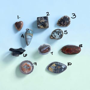 May include: Ten distinct rings featuring various gemstones set in metal, numbered one through ten. The collection includes heart-shaped, square, oval, and teardrop-cut stones in colors ranging from brown, black, and white to orange and blue-grey.