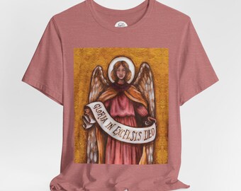 May include: A dusty rose t-shirt featuring a detailed painting of an angel holding a scroll. The angel has large wings and a halo, with the text "GLORIA IN EXCELSIS DEO" on the scroll. The background is a textured gold.