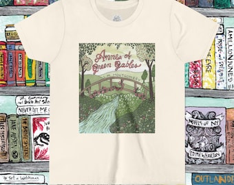 May include: Cream-colored t-shirt featuring a colorful illustration from the book "Anne of Green Gables" by L.M. Montgomery. The design includes a bridge, river, and trees. The shirt is displayed in front of a bookshelf filled with books.