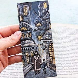 May include: Hand-painted bookmark featuring a detailed illustration of a cobblestone street with buildings and figures. The artwork is in shades of blue, gray, and yellow, with a dark night sky. The bookmark is held over an open book.