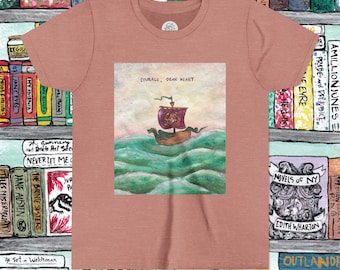 May include: A dusty rose t-shirt featuring a watercolor illustration of a ship sailing on green waves. The ship has a purple sail and the text "COURAGE, DEAR HEART" is above the image. The shirt is surrounded by a bookshelf.
