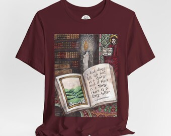 May include: Burgundy t-shirt featuring a detailed illustration of an open book with a landscape scene and text. The text reads "I had always felt life first as a story..." and "SOLI DEO GLORIA". The design includes a candle and a library.