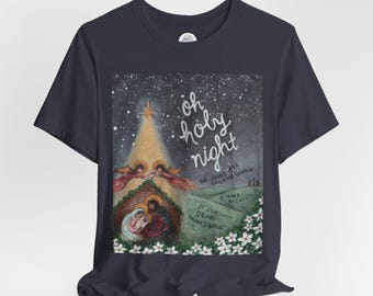 May include: Navy blue t-shirt featuring a Christmas scene with the text "oh holy night." The design includes a star, angels, and a nativity scene. White flowers are at the bottom of the image. The shirt is made of a soft material.