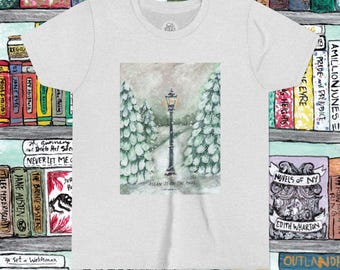 May include: A light gray t-shirt featuring a watercolor painting of a snowy path with a lamppost and evergreen trees. The text "ASLAN IS ON THE MOVE" is below the image. The shirt is surrounded by a bookshelf.