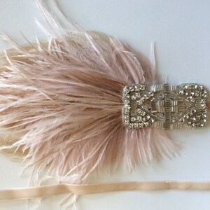 May include: A blush pink feather hair accessory featuring a rectangular silver-toned embellishment covered in clear crystals and metallic beads. The piece is attached to a thin, matching blush pink ribbon band that extends from both sides.