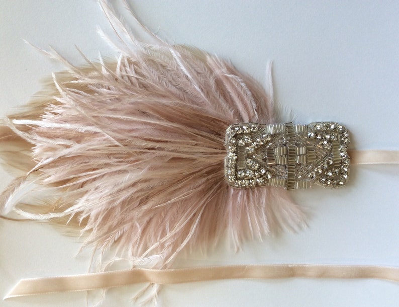May include: A blush pink feather hair accessory featuring a rectangular silver-toned embellishment covered in clear crystals and metallic beads. The piece is attached to a thin, matching blush pink ribbon band that extends from both sides.
