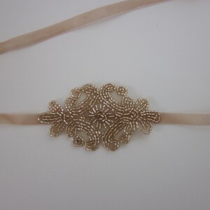 Peut inclure: Un bandeau de t&ecirc;te beige clair avec un motif floral central. Le motif floral est compos&eacute; de petites perles dor&eacute;es. Le bandeau est fait d'une mati&egrave;re douce et velout&eacute;e.