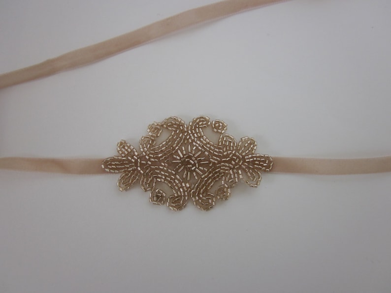 Peut inclure: Un bandeau de t&ecirc;te beige clair avec un motif floral central. Le motif floral est compos&eacute; de petites perles dor&eacute;es. Le bandeau est fait d'une mati&egrave;re douce et velout&eacute;e.
