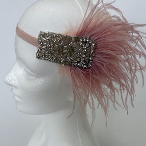 May include: A headband featuring a light pink elastic band and a rectangular decorative piece adorned with clear crystals and silver-toned beads. A cluster of soft, light pink ostrich feathers extends from the side of the beaded ornament.