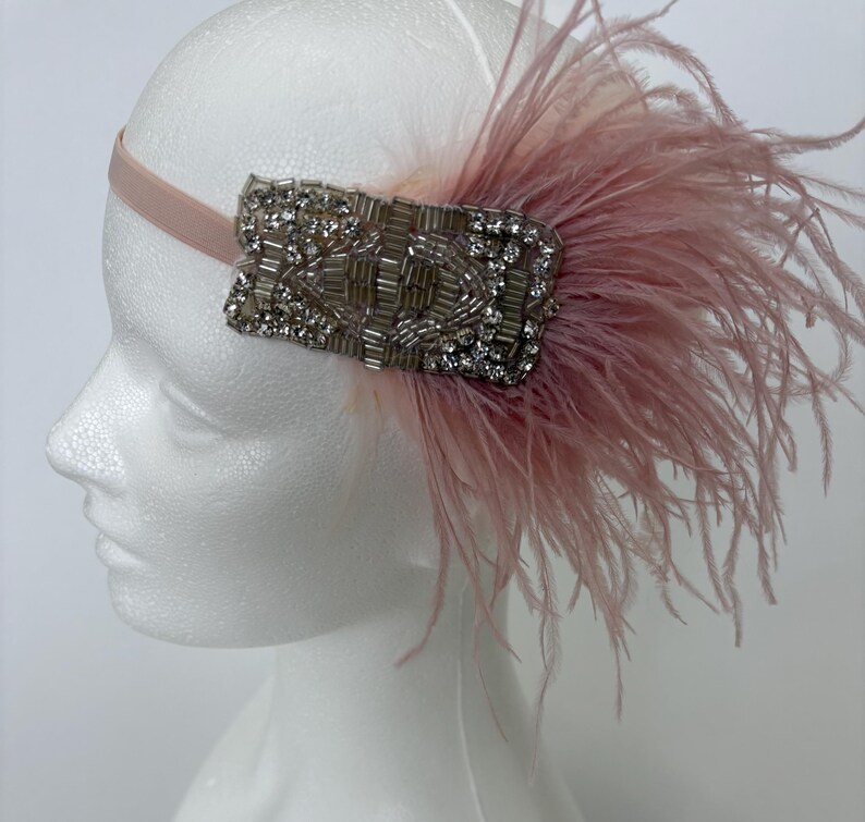 May include: A headband featuring a light pink elastic band and a rectangular decorative piece adorned with clear crystals and silver-toned beads. A cluster of soft, light pink ostrich feathers extends from the side of the beaded ornament.